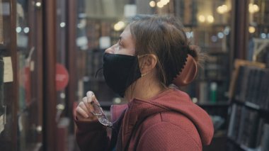 Gözlüklü ve maskeli ciddi bir beyaz kadın antika dükkanında eski bir kitaba benziyor.