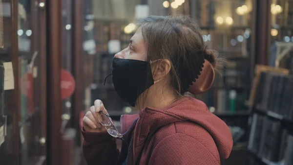 Serious caucasian woman in glasses and a mask looks old book in an antique store