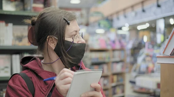 Woman in glasses and mask writes down names of books in the store in a notebook — Stock Photo, Image