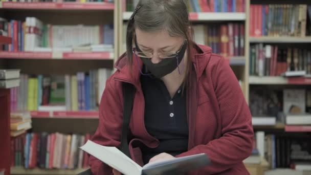 Caucasian woman wearing glasses and mask against virus reads a book in bookshop — Stock Video