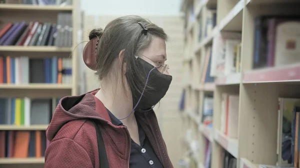Woman wearing glasses and mask against virus chooses book for reading in library
