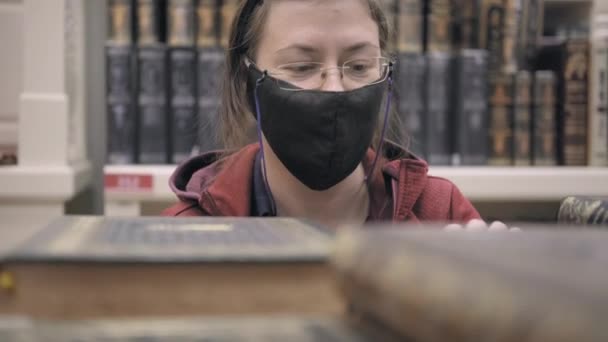 Femme blanche portant des lunettes et un masque est à la recherche d'un vieux livre dans la bibliothèque — Video