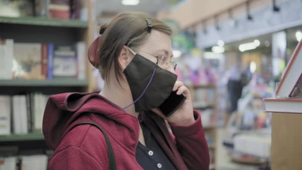 Mujer en una máscara contra el virus y gafas se para en la tienda y habla por teléfono — Vídeo de stock