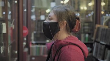 Gözlüklü ve maskeli ciddi bir beyaz kadın antika dükkanında eski bir kitaba benziyor.