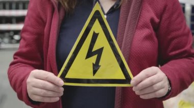 Kadının elinde üzerinde yıldırım işareti olan sarı üçgen bir tabela var. Yüksek volt