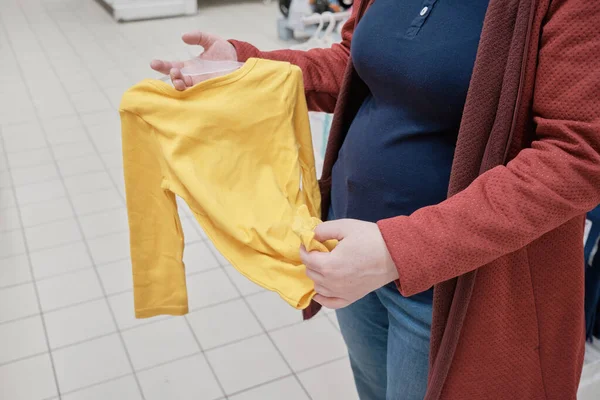 Kobieta w ciąży wybiera żółte ubrania - gorset dla dziecka w sklepie kupić — Zdjęcie stockowe