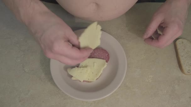 A kövér ember csinál egy szendvicset sajttal, kolbásszal és megtölti majonézzel, ketchuppal. — Stock videók