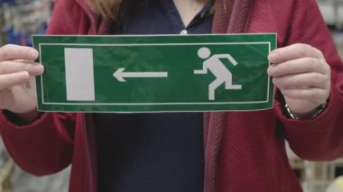 Kadının elinde acil çıkışı gösteren bir tabela var. Eller yakın çekim