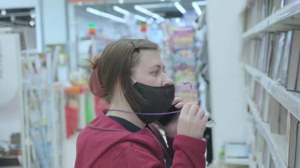 Žena v masce proti virům mluví po telefonu a vybere knihu v knihkupectví — Stock video