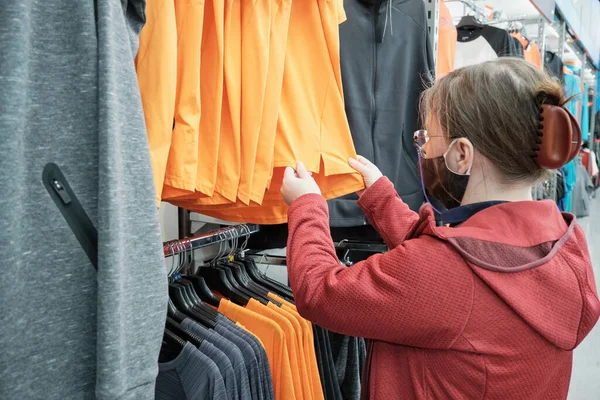 Mulher branca em máscara contra vírus escolhe uma camisa laranja para comprar em uma loja — Fotografia de Stock