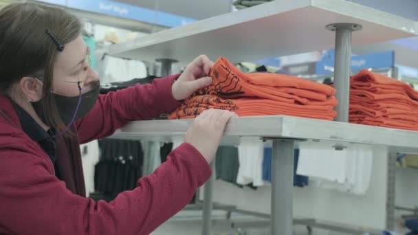 ウイルス対策マスクの女性は、購入する衣料品店でオレンジシャツを選択します — ストック動画