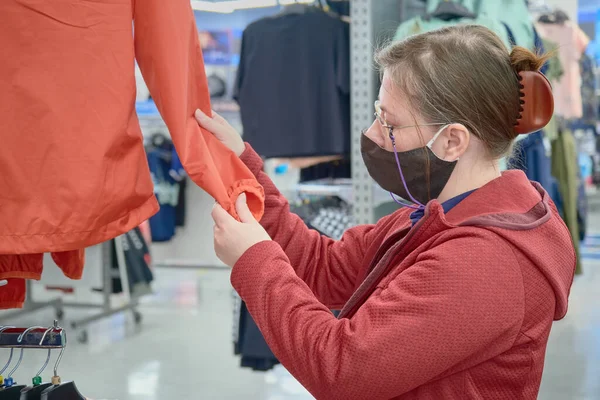 Mulher branca em máscara contra vírus escolhe um blusão vermelho para comprar na loja — Fotografia de Stock