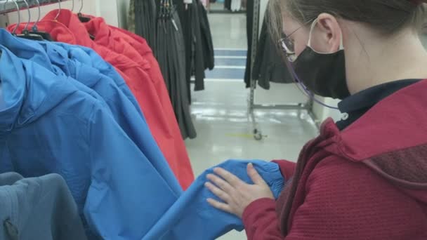 Mulher em máscara contra vírus escolhe uma jaqueta blusão azul na loja para comprar — Vídeo de Stock