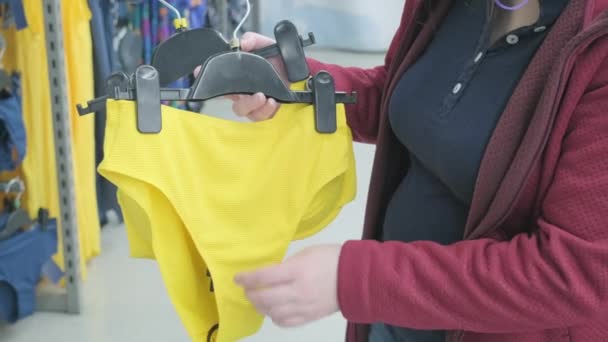 Кавказька вагітна жінка вибирає яскраво-жовтий, відкритий купальник в магазині, щоб купити — стокове відео