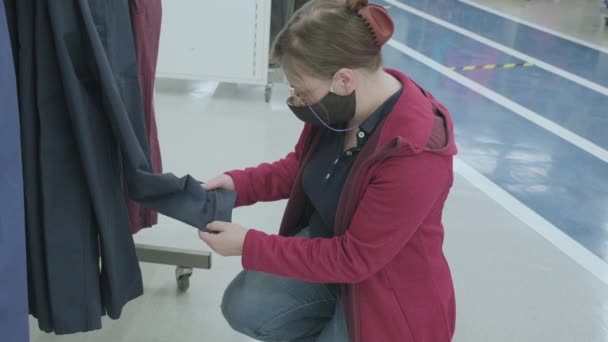 Vrouw met masker tegen virus kiest donkerblauwe broek in kledingwinkel te kopen — Stockvideo