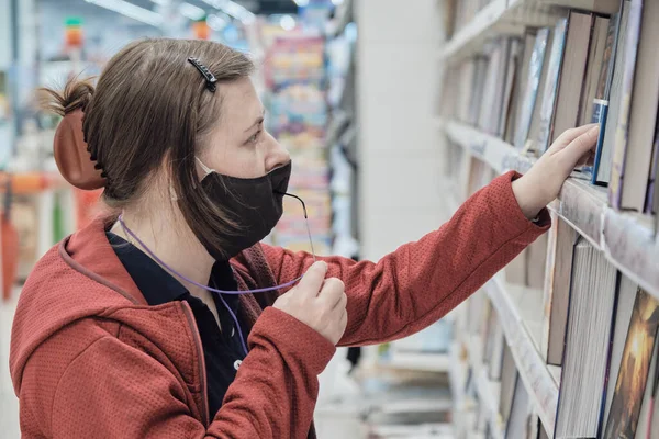 Woman wearing glasses and against virus mask picks out book from bookstore — Stock Photo, Image