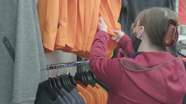 Mujer caucásica en máscara contra el virus elige una camisa naranja para comprar en una tienda — Vídeo de stock