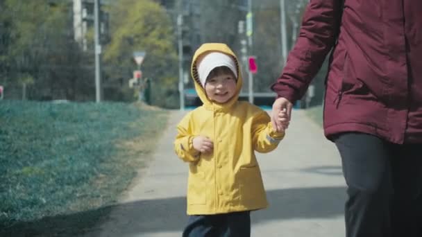 Дитина в жовтому плащі з капюшоном йде містом і тримає руку своєї матері — стокове відео