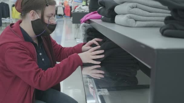 Mujer con máscara contra virus elige pantalones, sudaderas con capucha en tienda de ropa para comprar — Vídeos de Stock