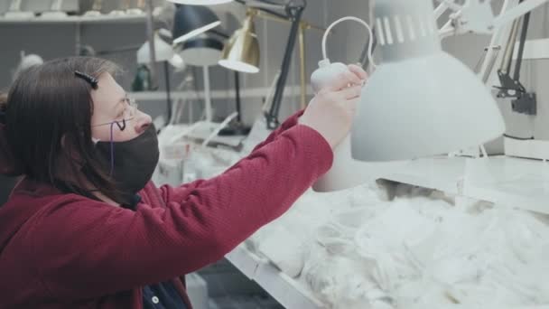 Blanke vrouw met masker tegen virus in een winkel kiest tafellamp te kopen — Stockvideo