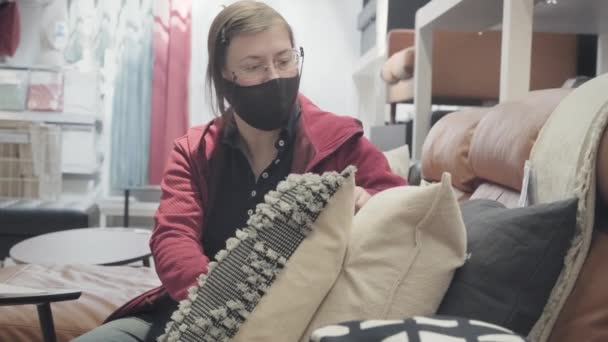 Mujer en contra de la máscara de virus se sienta en el sofá en la tienda y selecciona almohadas suaves — Vídeo de stock
