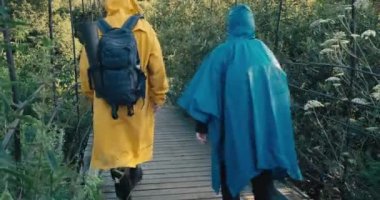 Man and woman hiker in raincoats walk on suspension bridge. Clear day. From back
