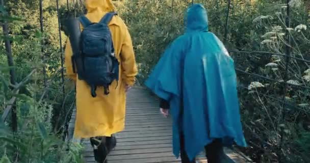 Man and woman hiker in raincoats walk on suspension bridge. Clear day. From back — Stockvideo