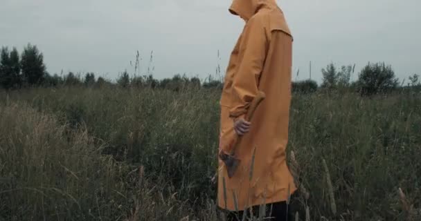 Man in raincoat and carrying axe is walking through field at dusk. From back — Vídeo de Stock