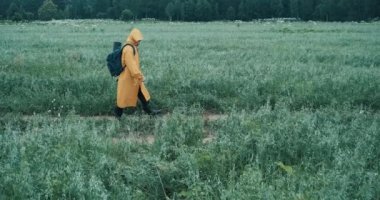 Man in a yellow raincoat and a backpack walks through a field with grass