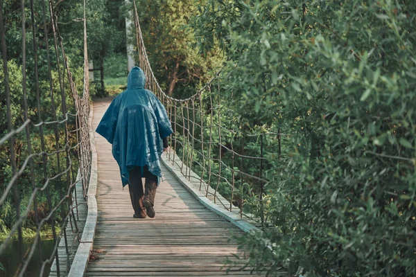 Girl with backpack under blue raincoat is engaged in hiking. Suspension bridge