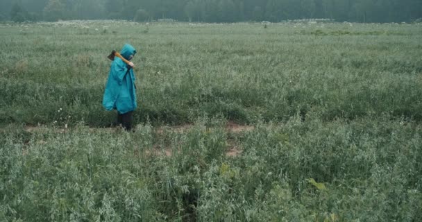 Woman in blue raincoat and carrying an axe is walking through the field at dusk — Stock Video