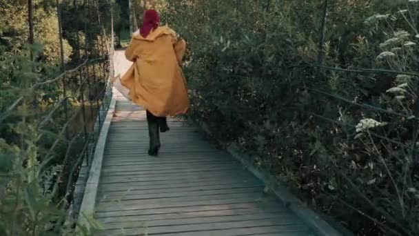 Man in een gele regenjas en een rode bandana loopt langs een houten hangbrug — Stockvideo