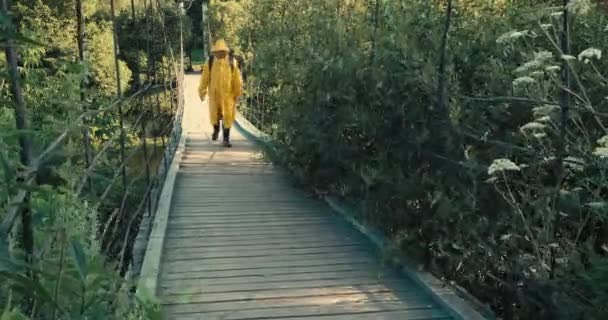 El hombre con mochila, en un impermeable amarillo, se dedica al senderismo. Puente colgante — Vídeo de stock