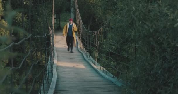 Чоловік пішохідний турист у жовтому плащі, що йде дерев'яним підвісним мостом — стокове відео