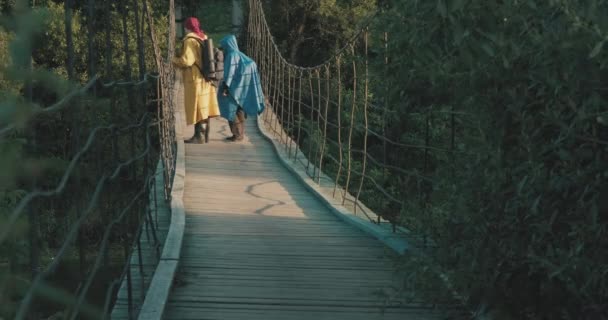 Caminhantes de homem e mulher em capas impermeáveis caminham na ponte suspensa. Pôr do sol — Vídeo de Stock
