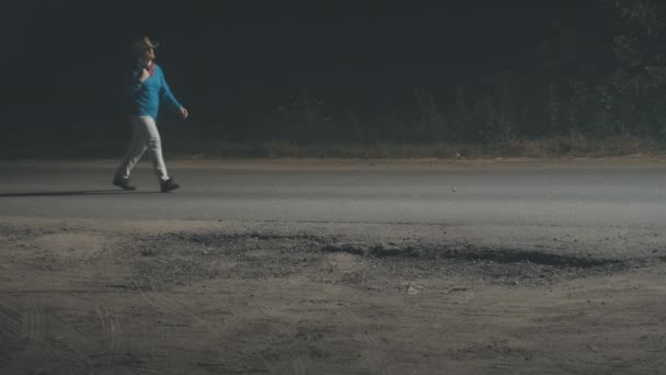 Hombre con sombrero de vaquero con una chaqueta sobre su hombro está caminando a lo largo de un camino nocturno — Vídeos de Stock