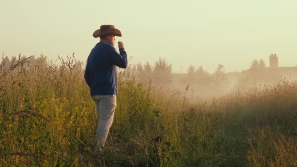 O homem de chapéu de cowboy está no campo, admira o sol do amanhecer. Homem mordisca lâmina de grama — Vídeo de Stock