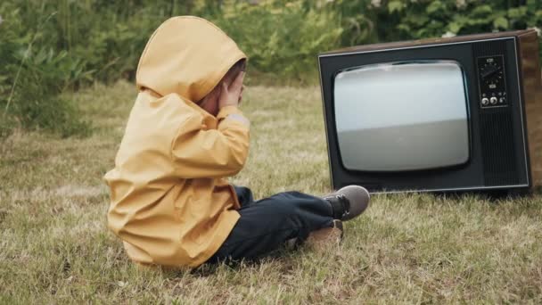 Un niño pequeño está mirando un viejo televisor y se cubre los ojos con sus palmas. — Vídeo de stock