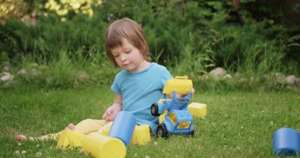 Pequena criança bonita está brincando com carro de brinquedo e cubos sentados na grama verde — Vídeo de Stock