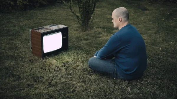 El hombre calvo está sentado en la hierba y mirando la vieja televisión retro. La TV está aturdida. Fotos de stock libres de derechos