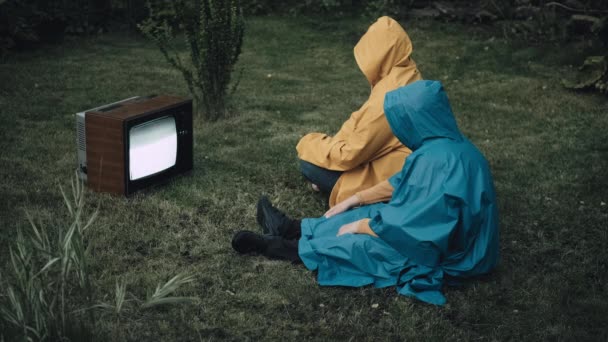 Homme et femme en imperméables avec capuche sont assis sur l'herbe et regarder une vieille télévision — Video