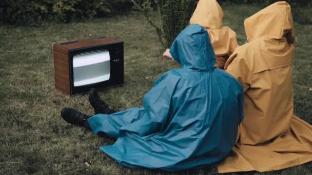 Tříčlenná rodina sedí na trávě v pláštěnkách a dívá se na starou retro televizi — Stock video