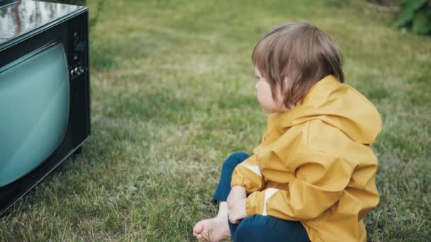 Small child in yellow jacket is sitting on grass and watching an old retro TV — Stock Video