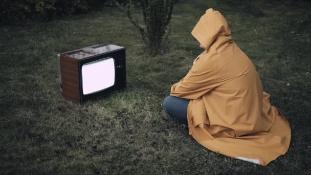 El hombre con un impermeable amarillo está sentado en la hierba y mirando la vieja televisión retro — Vídeos de Stock