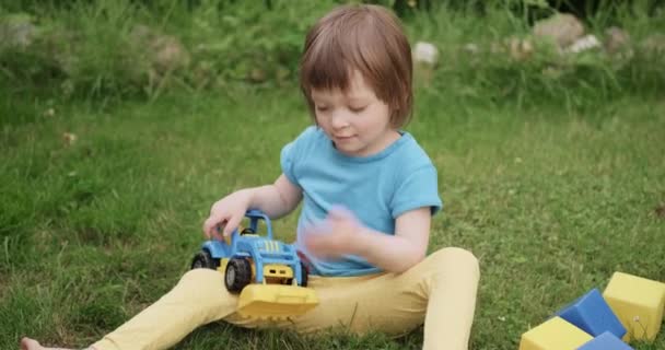 Pequena criança ruiva está sentada na grama e brincando com o carro de brinquedo de construção — Vídeo de Stock