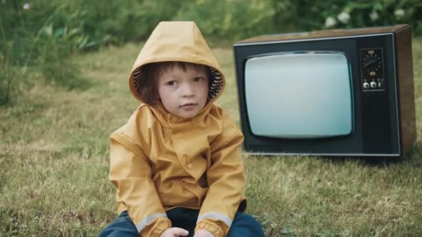 Triest kind gekleed in regenjas zit op gras. Achter hem, een oude retro TV. — Stockvideo