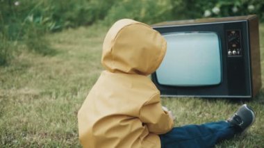 Yağmurluk giymiş bir çocuk retro TV izliyor. Sonra çocuk yükselir, televizyona dokunur.
