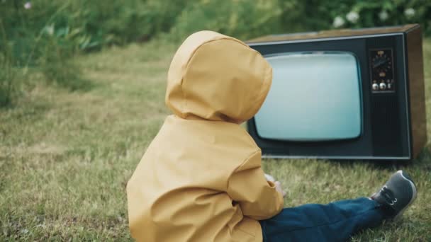 Dziecko ubrane w płaszcz przeciwdeszczowy ogląda telewizję retro. Potem dziecko się podnosi, dotyka telewizora. — Wideo stockowe