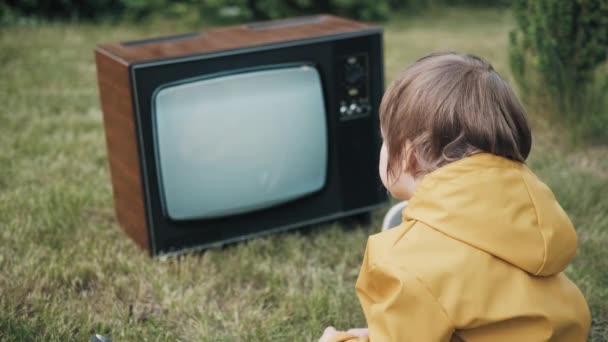 Kleines Kind sitzt auf Gras und sieht einen alten Retro-Fernseher. Fernsehen ist Strolch — Stockvideo