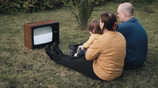 Семья с маленьким ребенком сидят на траве и смотрят старый ретро-телевизор — стоковое видео